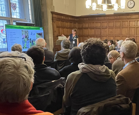 Speaker presenting to an audience in a wood-panelled conference room, with a presentation displayed on a large screen at the Real Oxford Farming Conference.