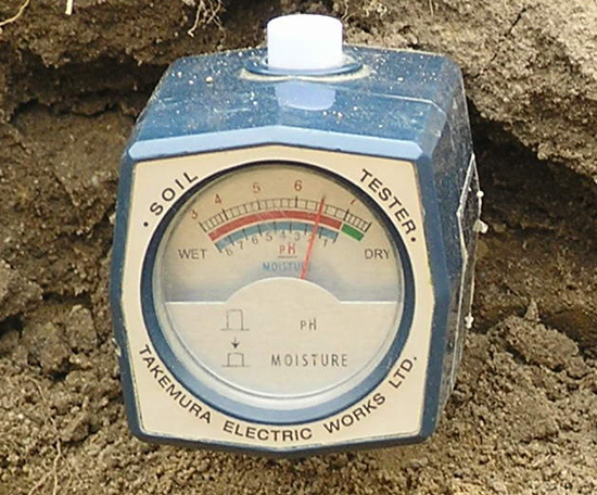  Soil pH and moisture meter inserted into soil, showing a measurement reading on an analogue dial.