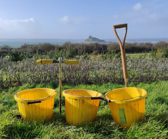 Soil sampling equipment, including buckets, a soil probe and a spade, arranged in a field overlooking a coastal landscape. 